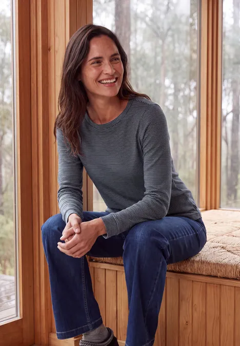 Langarm Shirt mit schmalem Streifenmuster urban dark blue Eine lächelnde Frau in grauem Pullover und Jeans sitzt auf einer Holzbank vor einem Fenster.