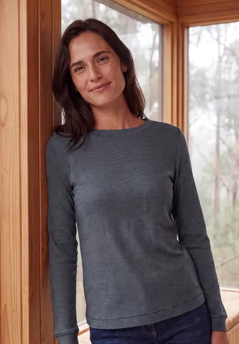 Langarm Shirt mit schmalem Streifenmuster urban dark blue Eine Frau in grauem Langarmshirt lehnt lächelnd in einem hellen Holzhaus an einem Türrahmen.