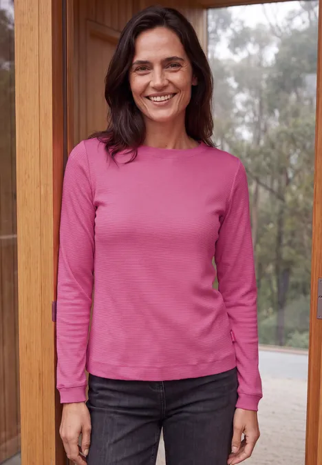 Langarm Shirt mit schmalem Streifenmuster pink flash Frau steht vor Holzrahmenfenster, trägt rosa Langarmshirt und schwarze Jeans, lächelt entspannt.