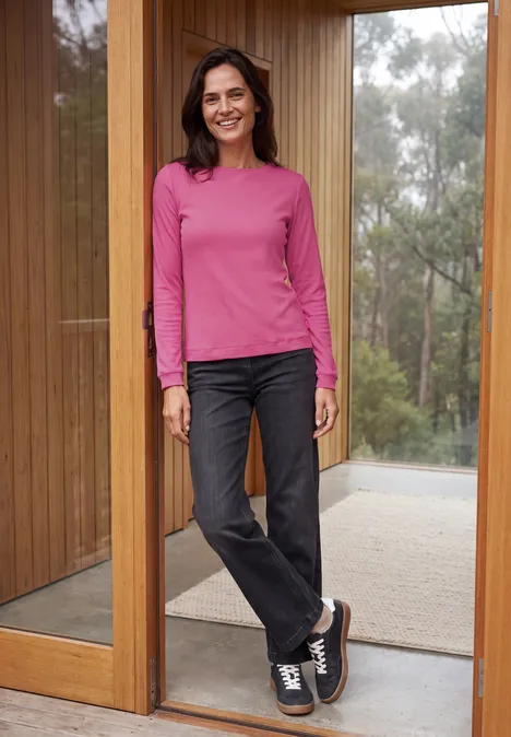 Langarm Shirt mit schmalem Streifenmuster pink flash Frau lehnt lächelnd im Türrahmen, trägt pinkes Oberteil, schwarze Jeans und Sneaker vor modernem Holzhauseingang