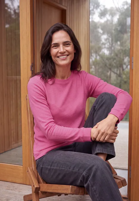 Langarm Shirt mit schmalem Streifenmuster pink flash Eine lächelnde Frau in einem pinken Pullover sitzt entspannt auf einem Holzstuhl vor großen Fenstern.