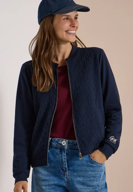 Jonge vrouw lacht, draagt een donkerblauw gewatteerd jasje, bordeaux shirt, spijkerbroek en pet.