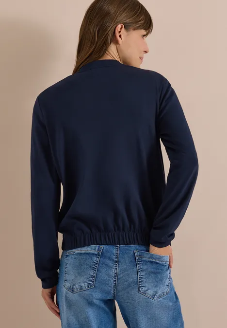 Rugaanzicht van een vrouw in een donkerblauw longsleeve met elastische zoom en blauwe jeans, handen in de zakken.