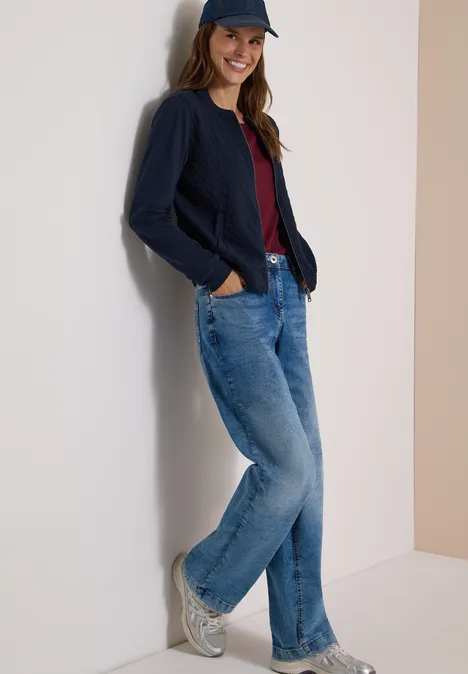 Vrouw leunt tegen een muur, draagt jeans, donkerblauwe cardigan, rood shirt, witte sneakers en pet.