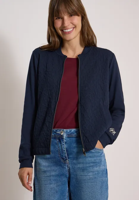 Jonge vrouw lacht, draagt een blauwe jas, een bordeauxrood shirt en een spijkerbroek.
