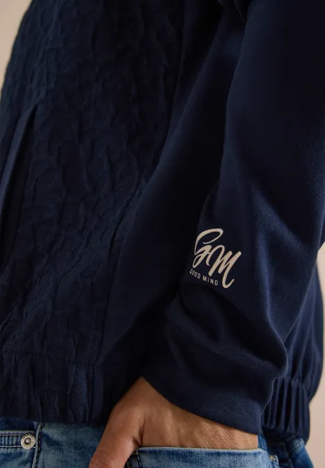 Close-up van de donkerblauwe doorgestikte mouw met goudkleurig HM-logo bij de manchetkant en hand in jeans