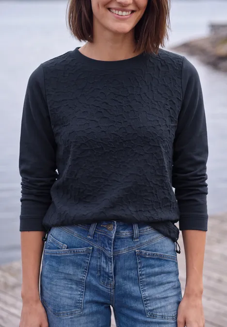 Shirt mit Boat Neck und Strukturmix urban dark blue Eine lächelnde Frau in schwarzem Pullover und blauer Jeans vor einer Wasserkulisse.