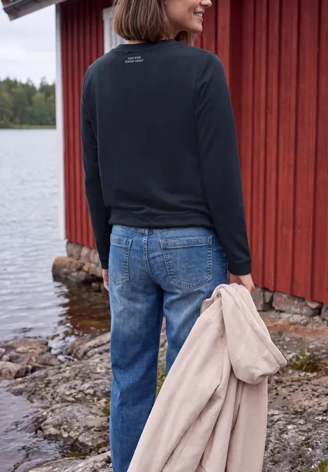 Shirt mit Boat Neck und Strukturmix urban dark blue Eine Frau steht in Jeans und Pullover vor einem roten Holzhaus am See und hält einen beigen Mantel.