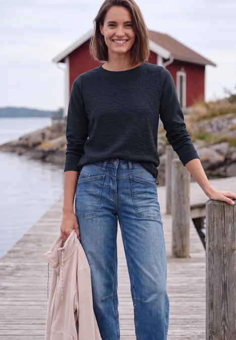 Shirt mit Boat Neck und Strukturmix urban dark blue Frau im schwarzen Pullover und Jeans steht auf einem Steg vor einem roten Haus am Wasser und hält eine Jacke.