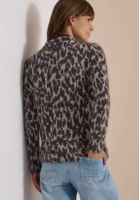 Vrouw draagt leopard gebreid vest en spijkerbroek, staand zijwaarts voor een lichte muur.