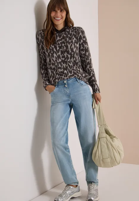 Vrouw in patroonblouse, blauwe spijkerbroek en tas, glimlachend tegen de muur geleund.