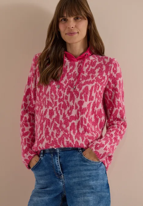 Vrouw draagt roze-witte capuchontrui met motief en blauwe jeans tegen een lichte achtergrond.