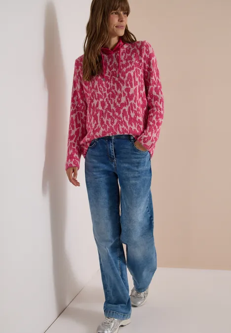 Vrouw staat bij een muur, draagt roze trui met print en losse blauwe jeans.