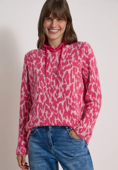 Vrouw lacht in een roze capuchontrui met patroon en jeans, op neutrale achtergrond.