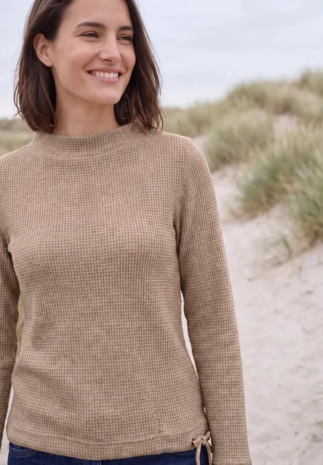 Shirt mit Funnel Neck und Waffelstruktur creamy mocha melange Eine Frau trägt einen beigen Pullover und lächelt in einer Dünenlandschaft.