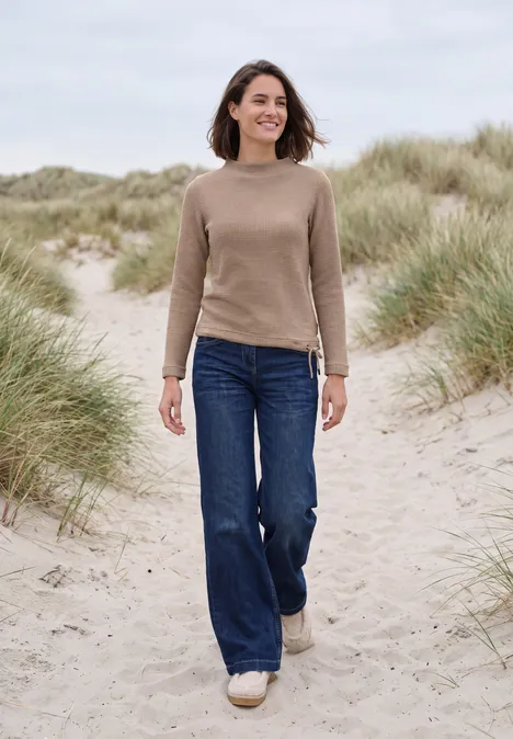 Shirt mit Funnel Neck und Waffelstruktur creamy mocha melange Eine Frau geht über grasbewachsene Dünen am Strand, trägt Pullover, Jeans und weiße Schuhe.