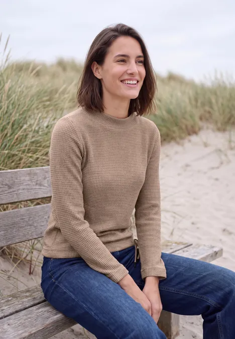 Shirt mit Funnel Neck und Waffelstruktur creamy mocha melange Frau sitzt lächelnd im Pullover und Jeans auf einer Bank am Strand mit Dünen im Hintergrund.