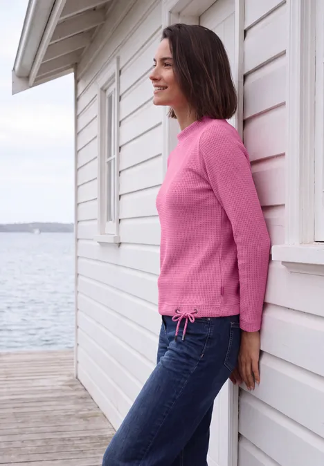 Shirt mit Funnel Neck und Waffelstruktur pink flash melange Frau in rosa Pullover lehnt an weißem Strandhaus, Blick auf ruhige See im Hintergrund.