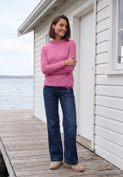 Shirt mit Funnel Neck und Waffelstruktur pink flash melange Frau in pinkem Pullover und Jeans steht auf Holzsteg vor weißem Haus am Wasser und lächelt.