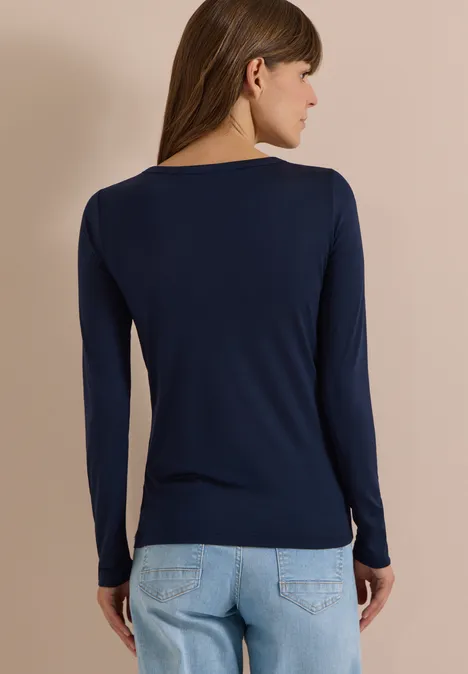 Frau von hinten in dunkelblauem Langarmshirt und hellen Jeans vor neutralem Hintergrund.