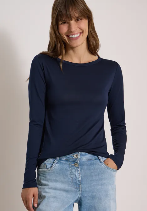 Eine Frau mit brünetten Haaren lächelt, sie trägt ein dunkelblaues Langarmshirt und Jeans.
