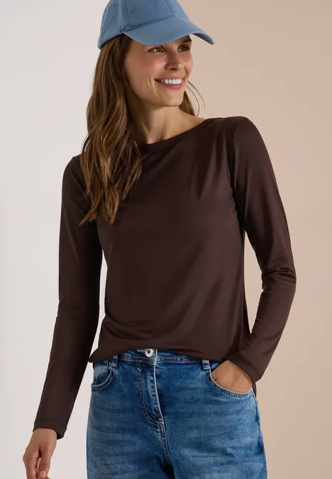 Junge Frau in braunem Langarmshirt, Jeans und Cap lächelt mit Händen in den Hosentaschen.