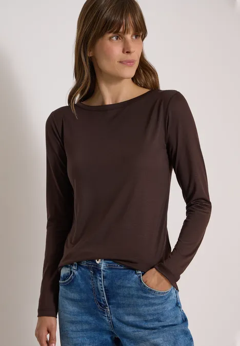 Junge Frau mit braunem Langarmshirt und Jeans, entspannt stehend und seitlich blickend vor hellem Hintergrund.