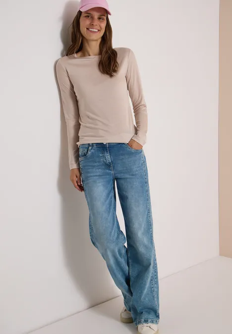Frau in beige Langarmshirt, blauen Jeans und rosa Cap lehnt lächelnd an der Wand.
