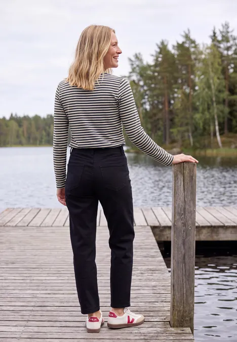 Frau in gestreiftem Pullover und schwarzer Hose steht auf einem Steg am See und blickt aufs Wasser.