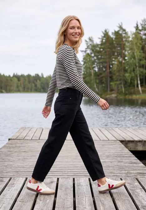 Eine fröhliche Frau in gestreiftem Shirt und schwarzen Hosen geht auf einem Holzsteg an einem See entlang.
