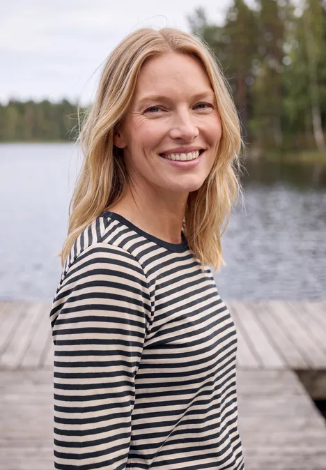Eine lächelnde blonde Frau in gestreiftem Shirt steht an einem Steg am See, umgeben von Wald.