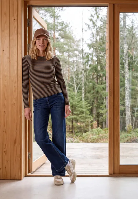 Shirt met lange mouwen, boothals en strepen truffle brown Een vrouw in een gestreept shirt, spijkerbroek en muts staat ontspannen in een houten ruimte voor grote ramen.