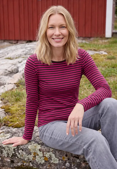 Shirt met lange mouwen, boothals en strepen pink flash Een glimlachende vrouw zit op een rots voor een rood houten huis.