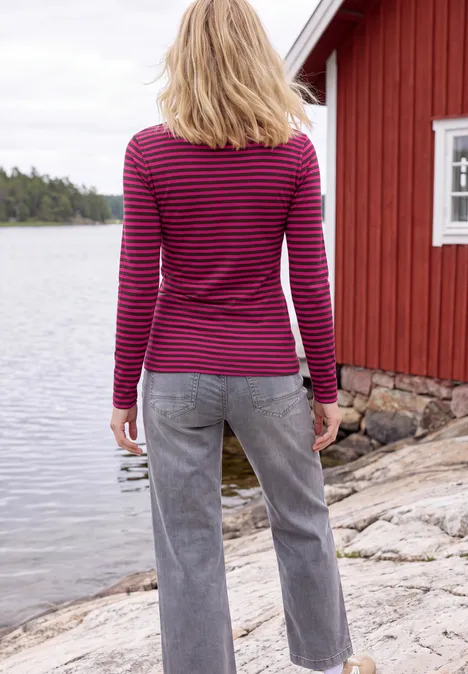 Shirt met lange mouwen, boothals en strepen pink flash Iemand in gestreept shirt en jeans staat op een rotsachtige oever voor een rood bootshuis.