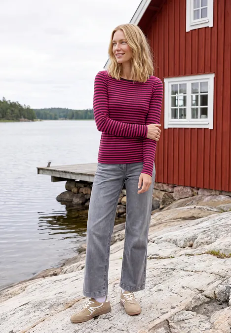 Shirt met lange mouwen, boothals en strepen pink flash Vrouw in gestreepte trui en grijze broek staat op rotsen voor een rood huis aan het meer.