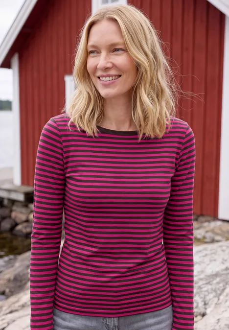 Shirt met lange mouwen, boothals en strepen pink flash Een lachende vrouw in een roze-paars gestreept shirt voor een rood houten huis aan het water.