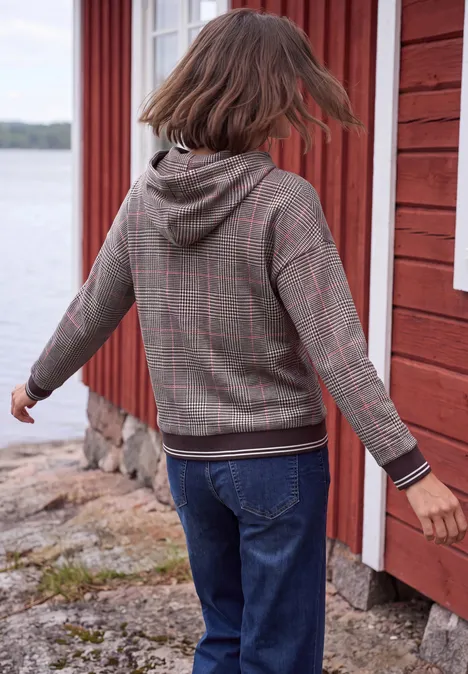 Eine Person in kariertem Hoodie und Jeans steht seitlich an einer roten Holzhütte am Wasser.