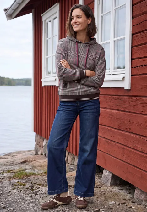 Eine lächelnde Frau in Jeans und Hoodie lehnt vor einem roten Holzhaus am See.