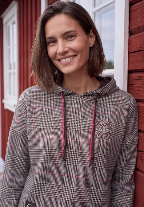 Lächelnde Frau vor rotem Holzhaus, trägt karierten Hoodie mit pinken Akzenten und Kordelzügen