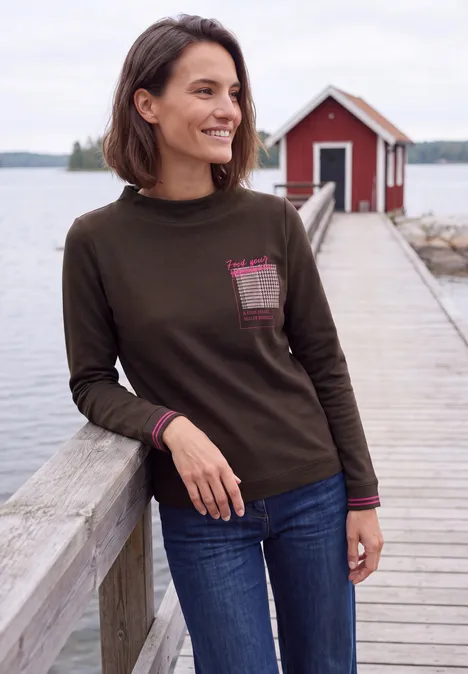 Langarm Shirt mit Funnel Neck und Print truffle brown Eine Frau lehnt lächelnd an einem Holzgeländer auf einem Steg vor einem roten Häuschen am See.