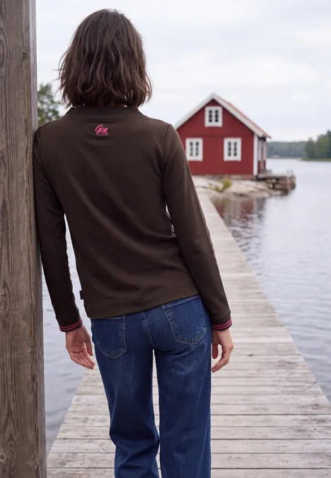 Langarm Shirt mit Funnel Neck und Print truffle brown Frau in Jeans und dunklem Pullover steht auf Holzsteg und schaut auf rote Hütte am ruhigen See.
