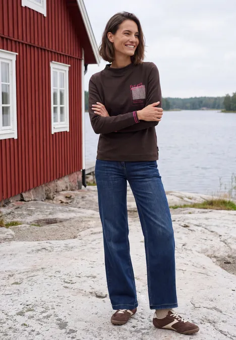 Langarm Shirt mit Funnel Neck und Print truffle brown Frau in braunem Pullover und blauen weiten Jeans steht lächelnd auf Felsen vor rotem Haus am See.