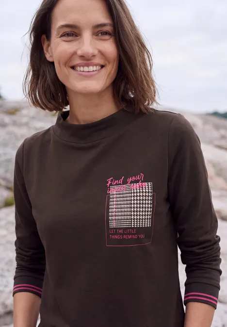 Langarm Shirt mit Funnel Neck und Print truffle brown Frau in dunkelgrünem Langarmshirt mit Print, lächelt vor felsiger Küstenkulisse.