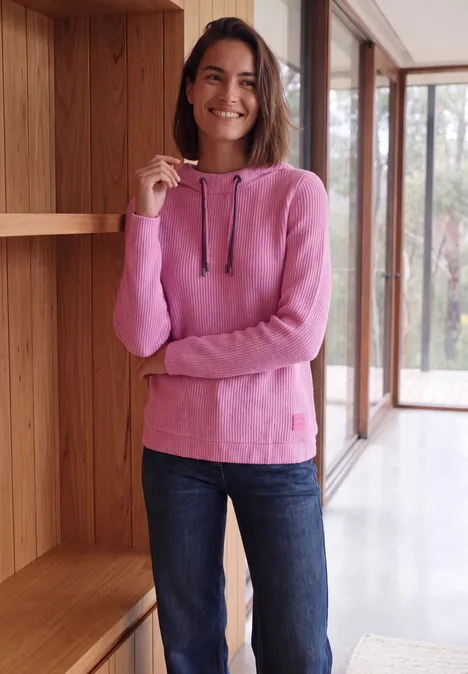 Lachende vrouw in een roze capuchontrui en spijkerbroek, staand bij het raam in een modern interieur.