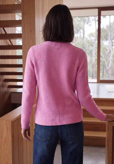 Vrouw van achteren in een roze trui en spijkerbroek, staand in een kamer met houten meubels.