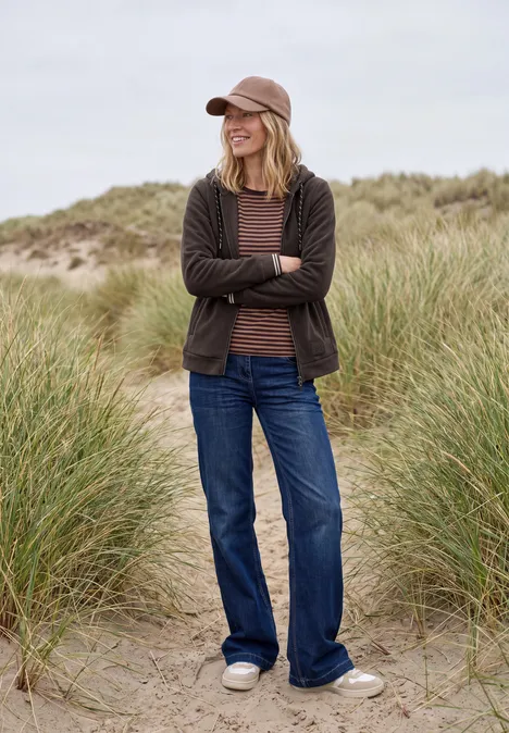 Langarm Fleece Jacke mit Kapuze truffle brown Eine Frau in Jeans, Cardigan und Cap steht lächelnd mit verschränkten Armen in grasbewachsenen Dünen.