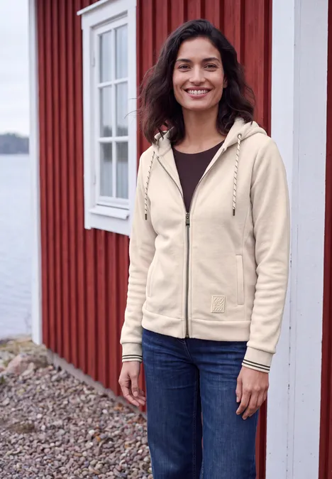 Langarm Fleece Jacke mit Kapuze cashmere beige Eine junge Frau in heller Jacke und Jeans steht lächelnd vor einem roten Holzhaus am Wasser.