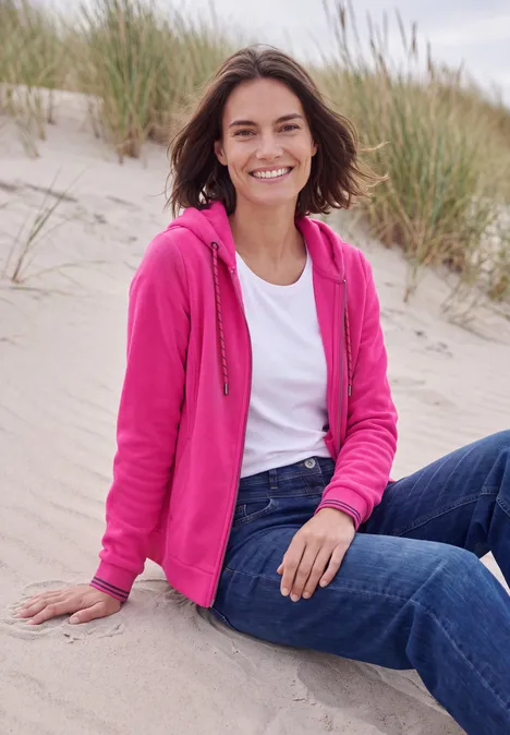 Langarm Fleece Jacke mit Kapuze pink flash Frau sitzt lächelnd im Sand vor Dünen, trägt pinke Jacke, weißes T-Shirt und Jeans.