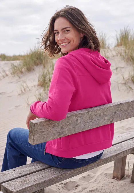Langarm Fleece Jacke mit Kapuze pink flash Eine lächelnde Frau in pinker Jacke sitzt auf einer Holbank am Dünenstrand.