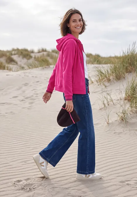 Langarm Fleece Jacke mit Kapuze pink flash Frau in pinkem Hoodie, blauen Jeans und weißen Sneakern spaziert lächelnd durch Dünen und hält einen Hut.